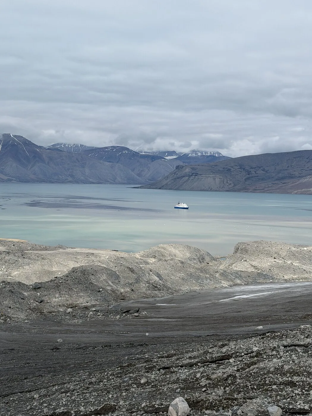 Grey moraine in the foreground, a glacial-milk fjord, an expedition ship at anchor for scale